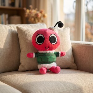 May include: A pink insect-shaped plush toy with large black eyes and a smiling mouth. It wears a green and white sweater with crosses and a white hat. The toy is on a beige sofa.