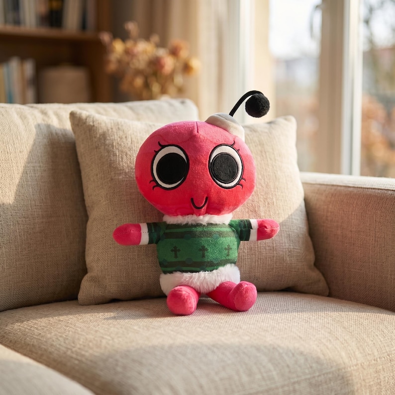 May include: A pink insect-shaped plush toy with large black eyes and a smiling mouth. It wears a green and white sweater with crosses and a white hat. The toy is on a beige sofa.