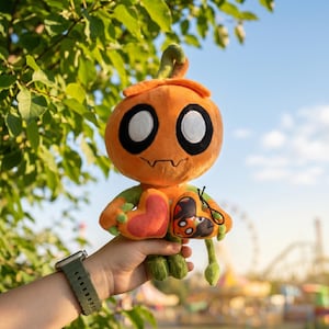 May include: An orange plush toy shaped like a pumpkin with large black eyes and a stitched mouth. The toy has green arms and legs, and a heart-shaped patch. The background is a sunny day with green foliage.