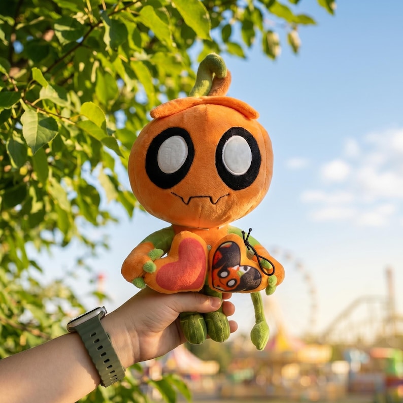 May include: An orange plush toy shaped like a pumpkin with large black eyes and a stitched mouth. The toy has green arms and legs, and a heart-shaped patch. The background is a sunny day with green foliage.