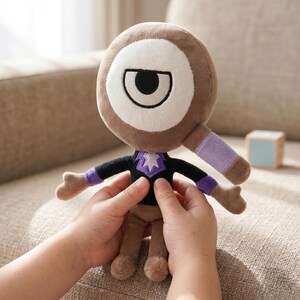 Puede incluir: Un peluche con una cabeza grande y redonda con un ojo blanco y un contorno marr&oacute;n. El juguete tiene una camisa negra con un dise&ntilde;o de estrella p&uacute;rpura, piernas marrones y detalles p&uacute;rpuras en los brazos. El juguete est&aacute; siendo sostenido por una persona.