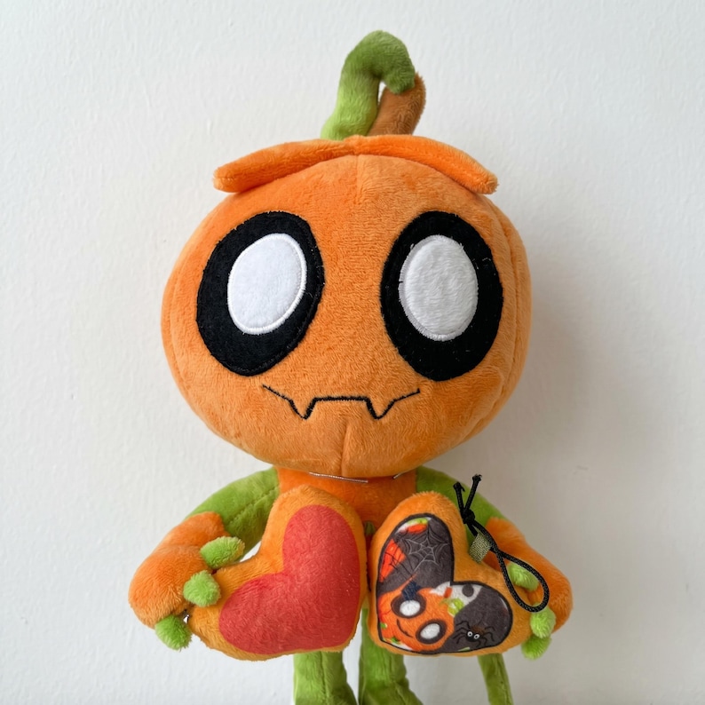 May include: Orange plush pumpkin figure with black eyes, a stitched mouth, and green stem and arms. The figure holds two heart-shaped accessories, one red and one with a Halloween design featuring a spider and jack-o'-lantern.