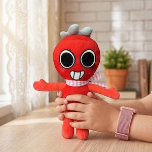 May include: A red plush toy with large black eyes, a white smile, and a pink and white scarf. The toy has small blue accents on top of its head and is being held by a person.