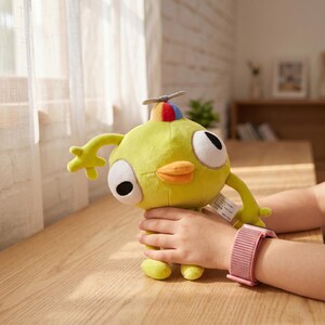 May include: A lime green plush toy with large, round eyes, an orange beak, and a propeller on its head. The toy is being held by a person wearing a pink watch. The toy has a whimsical design.