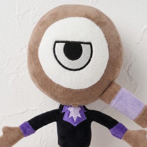 Puede incluir: Un peluche con una gran cabeza circular marr&oacute;n con un ojo blanco y una pupila negra. El juguete tiene un cuerpo negro con detalles morados en las mangas y el cuello. Los brazos est&aacute;n extendidos y el juguete tiene una apariencia amigable y de dibujos animados.