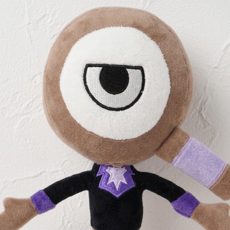 Puede incluir: Un peluche con una gran cabeza circular marr&oacute;n con un ojo blanco y una pupila negra. El juguete tiene un cuerpo negro con detalles morados en las mangas y el cuello. Los brazos est&aacute;n extendidos y el juguete tiene una apariencia amigable y de dibujos animados.