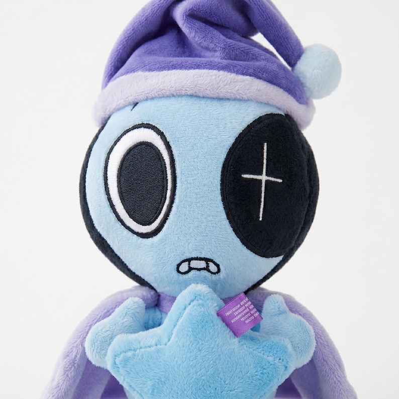 May include: A plush toy alien with a light blue face, large white-rimmed eyes, and a black patch over one eye. It wears a purple nightcap and holds a blue star. The toy has a stitched mouth and a purple cape.