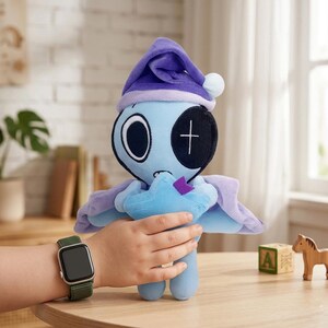 May include: A plush toy in the shape of a blue creature with a purple nightcap and cape. The toy has large eyes, a stitched cross on one eye, and a star-shaped object in its hands. The toy is being held by a child.