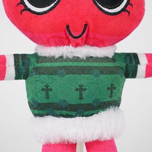 May include: A plush toy with a red head and pink limbs. It wears a green sweater with cross and dot patterns, trimmed with white faux fur. The toy has large black eyes and a curved smile.