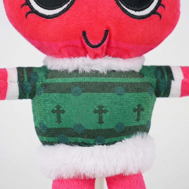 May include: A plush toy with a red head and pink limbs. It wears a green sweater with cross and dot patterns, trimmed with white faux fur. The toy has large black eyes and a curved smile.
