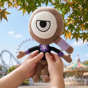 Puede incluir: Un peluche con un ojo grande y redondo y un cuerpo marr&oacute;n. El juguete tiene una camisa negra con un dise&ntilde;o de estrella morada y detalles morados en los brazos. El fondo incluye una noria y &aacute;rboles.