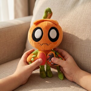 May include: An orange plush toy shaped like a pumpkin with large white eyes, black outlines, and a small, smiling mouth. The toy has green arms, legs, and a stem. A butterfly design is on the side. The toy is held by two hands.