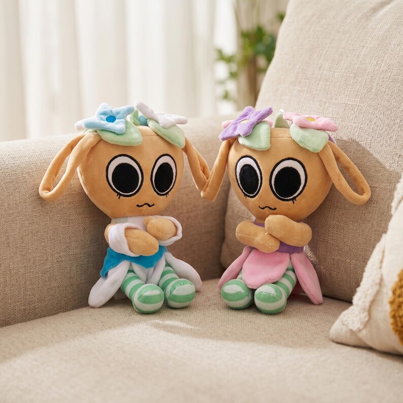 May include: Two plush dolls with large black eyes, tan fur, and long ears sit on a beige couch. Each doll wears a flower crown and has striped green and white legs. One doll has a blue and white outfit, and the other has a pink and purple outfit.