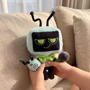 May include: A soft toy robot with a mint green square head, black screen, and green eyes. It has a green bow tie and black antennae. The robot is holding a smaller, matching toy.