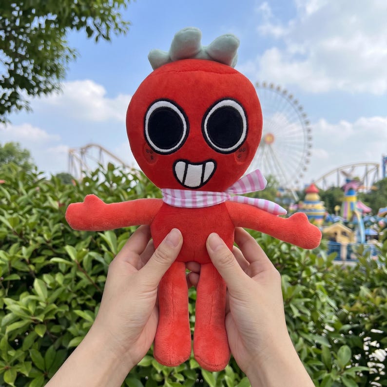 May include: A plush toy in the shape of a red character with large black eyes, a white mouth, and a green leafy top. It has a pink and white scarf and is being held up in front of a park background.