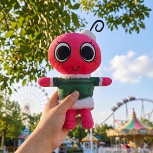 May include: A plush toy with a red head, large black eyes, and a small smile. It wears a green sweater with white trim and pink arms and legs. A black antenna curls from the top of its head. The toy is held in front of a blurred amusement park background.