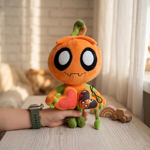 May include: An orange plush pumpkin character with large black and white eyes, a stitched mouth, and green arms and legs. It holds a red heart and a small pouch with a spooky design. The plush toy is designed for Halloween.