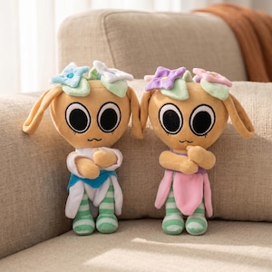 May include: Two plush dolls with large black eyes, tan bodies, and flower-shaped headbands. One doll wears a blue and white outfit, the other a pink and green outfit. Both have green and white striped legs and are posed on a beige couch.