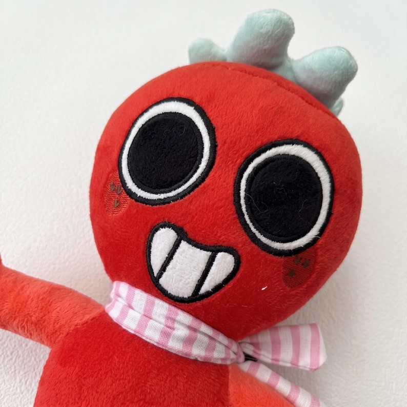 May include: A plush toy with a red head, large black eyes, and a smiling mouth. It has a light blue crown and a pink and white striped scarf. The toy is made of soft fabric.