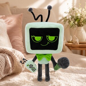 May include: A soft toy resembling a vintage television set, featuring a black screen and green trim. The toy has a green bow tie, black legs, and antennae. It holds a microphone and a remote control, all made from plush fabric.