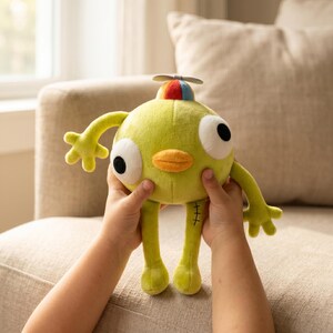 May include: A lime green plush toy with large black eyes, a yellow beak, and a small propeller on its head. The toy has arms and legs with stitched details. The toy is being held by a person.