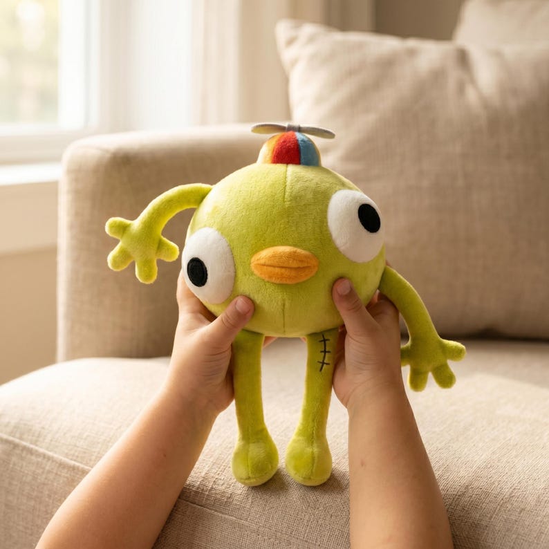May include: A lime green plush toy with large black eyes, a yellow beak, and a small propeller on its head. The toy has arms and legs with stitched details. The toy is being held by a person.