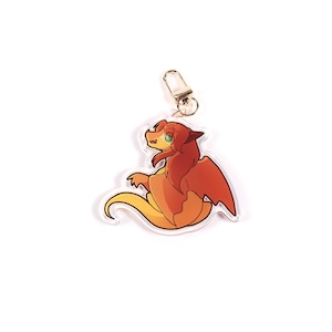 K&ouml;nnte beinhalten: Ein klarer Acryl-Schl&uuml;sselanh&auml;nger mit einem Cartoon-Drachen in Orange und Gelb. Der Drache hat rote Fl&uuml;gel und Haare sowie gr&uuml;ne Augen. Ein goldfarbener Verschluss ist oben am Schl&uuml;sselanh&auml;nger befestigt.