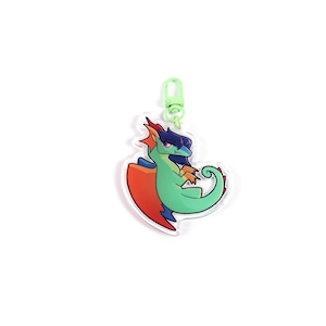 K&ouml;nnte beinhalten: Ein klarer Acryl-Schl&uuml;sselanh&auml;nger mit einem Cartoon-Drachen mit gr&uuml;nem K&ouml;rper, orangefarbenen und roten Fl&uuml;geln und lila Haaren. Der Drache ist an einem hellgr&uuml;nen Verschluss befestigt. Der Schl&uuml;sselanh&auml;nger befindet sich auf wei&szlig;em Hintergrund.