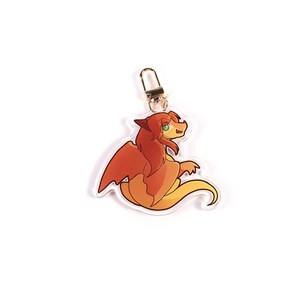 K&ouml;nnte beinhalten: Ein klarer Acryl-Anh&auml;nger mit einem Cartoon-Drachen in Orange- und Gelbt&ouml;nen. Der Drache hat einen freundlichen Ausdruck und ist an einem silberfarbenen Verschluss befestigt. Ein Accessoire mit Fantasy-Thema.