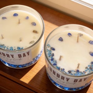 Puede incluir: Dos velas redondas de vidrio con cera blanca, cada una con tres mechas y piedras de cristal azul incrustadas en la superficie. Las etiquetas en los recipientes de vidrio transparente muestran el texto Laundry Day Scented Soy Candle en letras marrones.