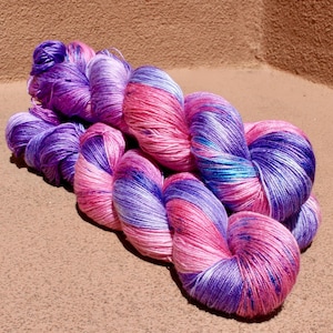 May include: A skein of hand-dyed yarn featuring a vibrant variegated color palette of bright pink, deep purple, and subtle hints of turquoise blue. The yarn has a smooth, lustrous sheen and is twisted into a compact, figure-eight shape.