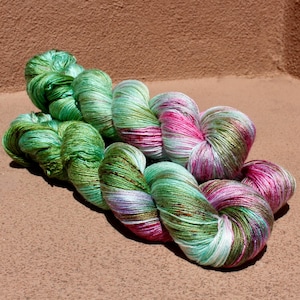May include: Two skeins of hand-dyed yarn featuring a variegated color palette of vibrant emerald green, soft mint, bright magenta, and subtle lavender tones. The yarn has a distinct sheen and a twisted, multi-ply texture throughout the fibers.
