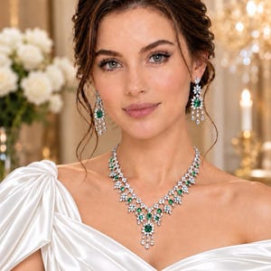 Puede incluir: Un conjunto de collar y pendientes colgantes en tono plateado con piedras de color verde esmeralda de corte ovalado rodeadas de cristales blancos brillantes. El collar presenta un diseño central con colgantes, a juego con los pendientes.
