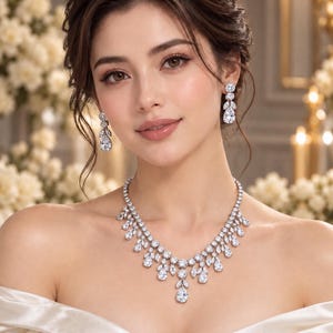 Può includere: Un set composto da collana e orecchini pendenti color argento con pietre trasparenti sfaccettate, a goccia e a taglio rotondo. La collana presenta una disposizione a cascata di pietre, mentre gli orecchini hanno una pietra rotonda superiore.