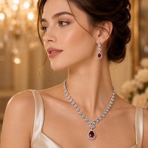 May include: A silver-toned necklace and matching drop earring set featuring pear-shaped deep red stones surrounded by a halo of small clear white crystals. The necklace has a central pendant design with a chain of circular crystal-encrusted links.