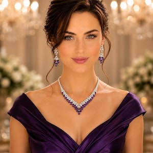 May include: A V-shaped necklace and matching drop earrings featuring marquise-cut purple gemstones and clear white stones set in silver-toned metal. The jewelry set is worn with a dark purple satin dress with a draped off-the-shoulder neckline.