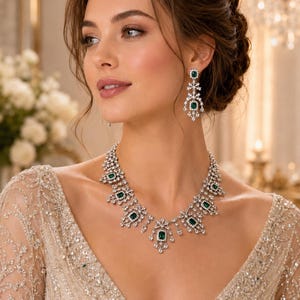 Può includere: Un set composto da collana e orecchini pendenti color argento con pietre verdi taglio smeraldo circondate da cristalli pavé trasparenti. La collana presenta un design centrale con segmenti multipli collegati e gli orecchini hanno un pendente verticale.