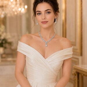 Puede incluir: Un vestido color marfil con hombros descubiertos que presenta un corpiño plisado con escote cruzado. El conjunto se complementa con un collar plateado y pendientes a juego, ambos engastados con piedras transparentes en forma de lágrima.