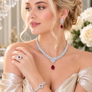 May include: A jewellery set consisting of a necklace, earrings, a bracelet, and a ring. Each piece features clear, round-cut stones set in silver-toned metal, accented with deep red, pear-shaped stones as the central focal points of the design.