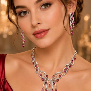 May include: A jewelry set consisting of a necklace and matching drop earrings. The pieces feature a silver-toned metal base set with numerous small clear stones and multiple teardrop-shaped deep red stones arranged in a flowing, vine-like pattern.