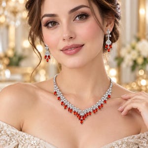 Puede incluir: Un conjunto de joyería compuesto por un collar y pendientes a juego. El collar presenta un diseño en forma de V con piedras rojas de talla marquesa y piedras redondas transparentes engastadas en metal de tono plateado.