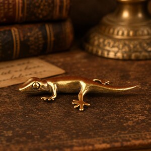 May include: A small, metallic gold-colored lizard figurine with a smooth, polished finish. The lizard is depicted in a crawling position with its four legs extended outward and its long, slender tail trailing behind it on the surface.