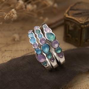May include: A set of three silver rings featuring an organic, wavy band design. Each ring is inlaid with multiple small, smooth, rounded gemstones in varying shades of translucent blue, teal, and soft purple, arranged in a cluster pattern.