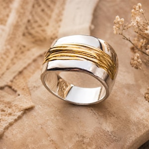 May include: A wide band ring featuring a polished silver finish with a textured, gold-toned wire wrap detail encircling the center. The ring has a smooth, reflective interior surface and a slightly irregular, organic shape along the outer edges.