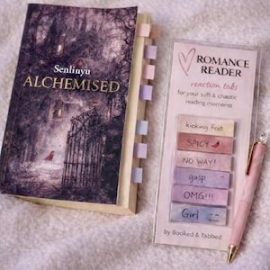 May include: A book titled "ALCHEMISED" with colorful tabs, a "ROMANCE READER" reaction tabs set, and a pink pen. The book cover features a dark, atmospheric illustration with a gate. The reaction tabs include phrases like "SPICY" and "OMG!!!".