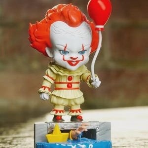 May include: A stylized vinyl figure of a clown with bright orange hair, white face paint, and red facial markings, wearing a cream-colored ruffled outfit with red buttons. The figure holds a red balloon and stands on a base featuring a sewer drain, a yellow paper boat, and a hand reaching out from blue water.