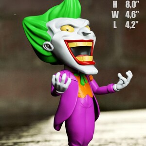 May include: A stylized Joker figurine with bright green hair, a white face, and a wide red-lipped grin. The character wears a purple suit jacket over an orange vest and green tie. Text indicates model dimensions of H 8.0 inches, W 4.6 inches, L 4.2 inches.
