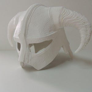 May include: A white, unpainted horned helmet featuring a central ridge, wide eye openings, and two large, curved horns extending from the sides. The surface has a textured, matte finish with visible molded details across the entire structure.