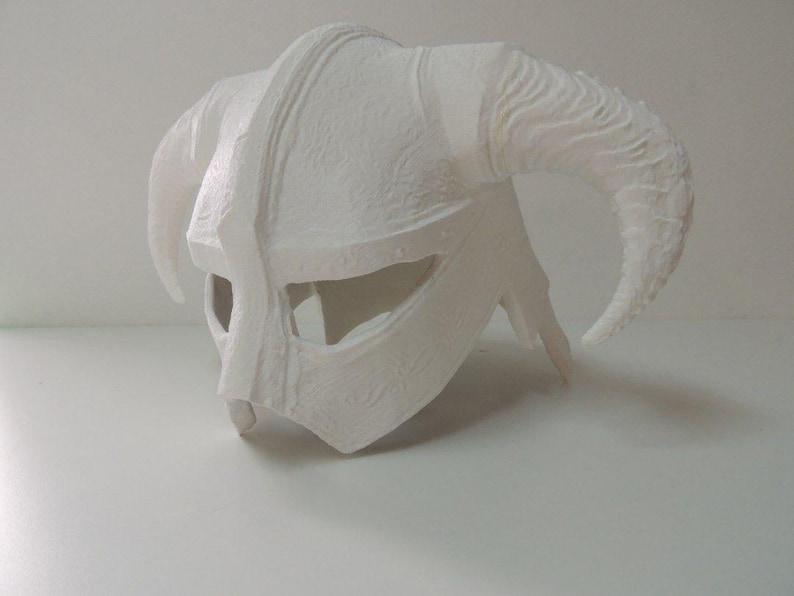May include: A white, unpainted horned helmet featuring a central ridge, wide eye openings, and two large, curved horns extending from the sides. The surface has a textured, matte finish with visible molded details across the entire structure.