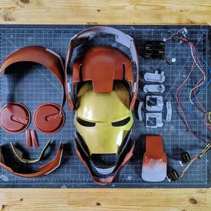 May include: A disassembled red and gold helmet kit featuring a central faceplate, various curved outer shell components, internal mounting brackets, a battery pack, and electronic wiring components laid out on a dark grid cutting mat.